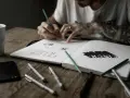 Un artiste auteur ou micro entrepreneur dessine dans son carnet, entouré de stylos, crayons et d'une tasse.