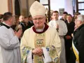 Uśmiechnięty biskup w stroju pontyfikalnym, otoczony przez wiernych i duchownych. W tle widać fragmenty kościoła, a na pierwszym planie szczegóły szat.