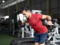 Man utför hantelflyes stående i ett gym. Han böjer sig framåt med rak rygg och lyfter hantlarna utåt med lätt böjda armbågar.