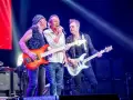 Trzech muzyków na scenie w głębokim fioletowym świetle Atlas Areny. Jeden śpiewa, dwaj grają na gitarach.