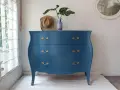 Cómoda antigua con **muebles pintados en azul** y tiradores dorados, decorada con un sombrero y un jarrón con hojas.