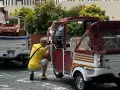 Turisti scattano foto a veicoli atipici, un Piaggio Ape e un tuk-tuk, in una strada soleggiata.