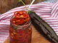 En burk med picklad chili, med en sked som lyfter upp en skiva. En mörk chili ligger bredvid.