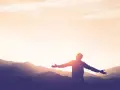 Silhouette einer Person mit ausgebreiteten Armen vor einer Bergkulisse bei Sonnenuntergang. Ein Symbol für Kraft und Hoffnung nach einer Krankheit.