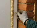 Ręka w rękawicy aplikuje piankę montażową do szczeliny między styropianem a ceglaną ścianą, zapewniając izolację.