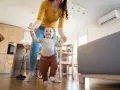 Mamá ayuda a su bebé a dar sus primeros pasos en una habitación cómoda, con el aire acondicionado bebes funcionando para mantener la temperatura ideal.