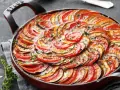 Ein wunderschönes Ratatouille-Rezept, das in einer roten Pfanne mit geschichteten Tomaten-, Zucchini- und Auberginenscheiben und frischen Kräutern präsentiert wird.