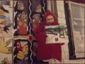 Maud Lewis, klädd i rött, håller upp en av sina charmiga vintermålningar framför sitt färgstarka hem.