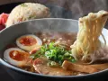 Un humeante tazón de ramen con huevo cocido, cebolleta y cerdo. Los palillos levantan fideos, listos para disfrutar de esta deliciosa receta ramen.