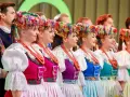 Szła dzieweczka w tradycyjnym stroju, z barwnym wiankiem na głowie, obok innych w ludowych strojach.