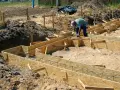 Budowa fundamentów: pracownik układa zbrojenie w drewnianym szalunku. Widać wiele desek na szalunek fundamentu, gotowych do zalania betonem.
