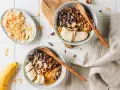 Due ciotole con granola fatta in casa, banana a fette, cioccolato fondente e burro d'arachidi. Cocco in scaglie a lato.