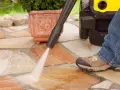 Pulire pavimento esterno con idropulitrice. Un getto d'acqua potente rimuove lo sporco dalle pietre del patio, mentre una persona in jeans e scarponi osserva.