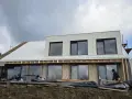 Trwa **wymiana blachy na dachu** nowoczesnego domu. Pracownicy montują nowe pokrycie, a budynek ma już zamontowane okna.