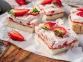 Saftige Stücke eines Erdbeerkuchen Blechs mit frischen Erdbeeren und Sahne, auf Backpapier angerichtet.