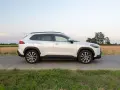 Biała Toyota Corolla Cross Hybrid stoi na poboczu drogi, otoczona polami i zieloną trawą.