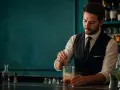 Barista esperto prepara un cocktail, con la sua paga base che riflette la sua abilità. Dietro di lui, bottiglie di liquori e un'atmosfera elegante.
