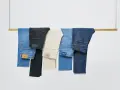 Cinco pares de pantalones vaqueros de distintos tonos cuelgan de una barra de madera. El **denim material** se presenta en azul claro, negro, beige, azul medio y azul oscuro.