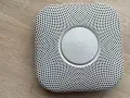 En vit Google Nest Protect-rökdetektor med ett runt "nest"-märke i mitten, placerad på ett trägolv.