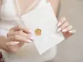 Dłonie z różowym lakierem trzymają białą kopertę zapieczętowaną złotą pieczęcią. Zastanawiasz się, ile za talerzyk na wesele?