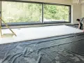 Pracownik układa płyty styropianowe do ocieplenie stropu betonowego. Duże okno wychodzi na zieleń.