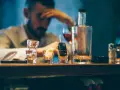 Ein Mann mit Kopfschmerzen sitzt am Tresen, neben Autoschlüsseln in einem Schnapsglas. Alkohol am Steuer unter 21 ist eine Gefahr.
