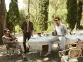 Uomini eleganti in un giardino, con un uomo seduto che indossa un completo marrone e un altro in piedi con un completo grigio. Il pantalone, in entrambi i casi, definisce lo stile.