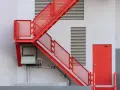 Nowoczesna, czerwona zabudowa schodów zewnętrznych z metalowymi stopniami i ażurowymi barierkami, prowadząca do czerwonych drzwi.