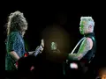 James Hetfield, z charakterystycznym wąsem i wytatuowanymi ramionami, gra na gitarze. Obok niego drugi muzyk z burzą włosów.