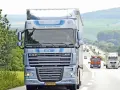 Niebieska ciężarówka DAF XF 105 na autostradzie, otoczona zielenią. W tle inne pojazdy i pagórkowaty krajobraz.