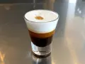 Kawa caffe coretto w szklance, z warstwami ciemnego espresso i białej pianki, ozdobiona złotą kropką.