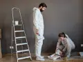 Pareja aprendiendo a pintar su hogar. Él sostiene un rodillo, ella vierte pintura. Hay cajas y una escalera.