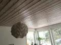 En vit lampskärm hänger från ett tak av träpanel. En tunn sladd dras i taket för att ansluta lampan.