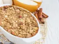 Pyszna, pieczona owsianka z jabłkiem i migdałami w białej kokilce, obok przekrojone gruszki i laski cynamonu.