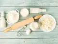 Zutaten für butter ersatz backen: Mehl, Milch, Quark, Ei, Schneebesen und Nudelholz auf blauem Holzuntergrund.