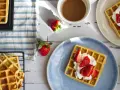 Frische belgische Waffeln mit Erdbeeren und Sahne auf einem blauen Teller, daneben eine Tasse Kaffee und weitere Waffeln auf einem Gitter.