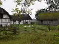 Stare chaty z krytymi strzechą dachami, gęsi pasące się na trawie. W tle drewniany płot i drzewa. Kluki cennik.