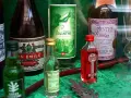 Butelki z absyntem: zielony, czerwony i ciemny. W tle szkło do absyntu i inne butelki. Absynt z czego? Z ziół i roślin.