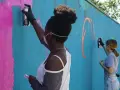 Ungdomar målar graffiti på en blå vägg. En flicka med afrohår sprayar rosa färg.