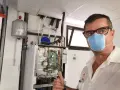 Técnico con mascarilla realiza mantenimiento aerotermia. Equipo abierto, circuito visible. Pulgar arriba.