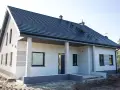 Nowy dom z szarą dachówką i jasną elewacją, ozdobioną subtelnymi, poziomymi pasami. Trwają prace nad malowaniem elewacji, nadając budynkowi nowoczesny wygląd.