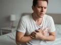 Mann hält sich die Brust, leidet unter Brustbein Schmerzen. Er sitzt auf einem Bett, sein Gesichtsausdruck ist besorgt.