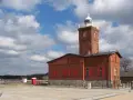 Ceglana latarnia morska Darłowo z czerwonymi okiennicami, na tle błękitnego nieba z chmurami.