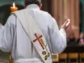 Ksiądz w białej szacie z krzyżem i kłosem na stułę, z rękami wzniesionymi w geście modlitwy, tworzy atmosferę religijnej suspensy.