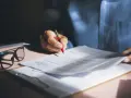 Persona revisa y firma una nota de encargo inmobiliaria, con gafas y bolígrafo rojo sobre documentos.