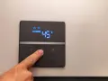 Dito, un dito preme il pulsante di un pannello digitale che mostra l'indicatore temperatura scaldabagno a 45°C, con icone Wi-Fi e di riscaldamento attive.