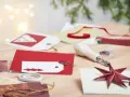 Förberedelser för att måla julkort. Rött glitterpapper, vita etiketter med julgransmotiv och en stjärnformad gemklämma ligger på ett träbord.