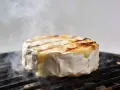 Un camembert au barbecue, fondant et fumant, cuit sur une grille. La croûte blanche se craquelle, révélant un cœur doré et coulant.
