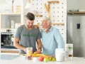 Två män i ett kök förbereder en smoothie med frukt. En burk med vegetariskt protein står på bänken.