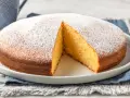 Torta soffice allo yogurt greco, cosparsa di zucchero a velo, con una fetta tagliata che ne rivela l'interno dorato.